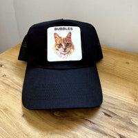 Front view of a black trucker cap with a custom cat portrait patch